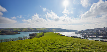 Snapes Point headland This is a landscape photograph of Snapes Point headland in Devon, England, United Kingdom, taken during the afternoon in early spring. The image prominently features the green grassy fields of Snapes Point extending towards the estuary, with the water of the Kingsbridge Estuary clearly visible. In the background on the right, the town of Salcombe can be seen nestled along the estuary’s edge, with its cluster of buildings adding detail to the coastal scene. The sky is partly cloudy with the sun high, casting bright light across the landscape, emphasizing the fresh spring vegetation on the headland. The photograph captures the characteristic countryside of Devon with rolling hills and waterways under clear, seasonal lighting.
